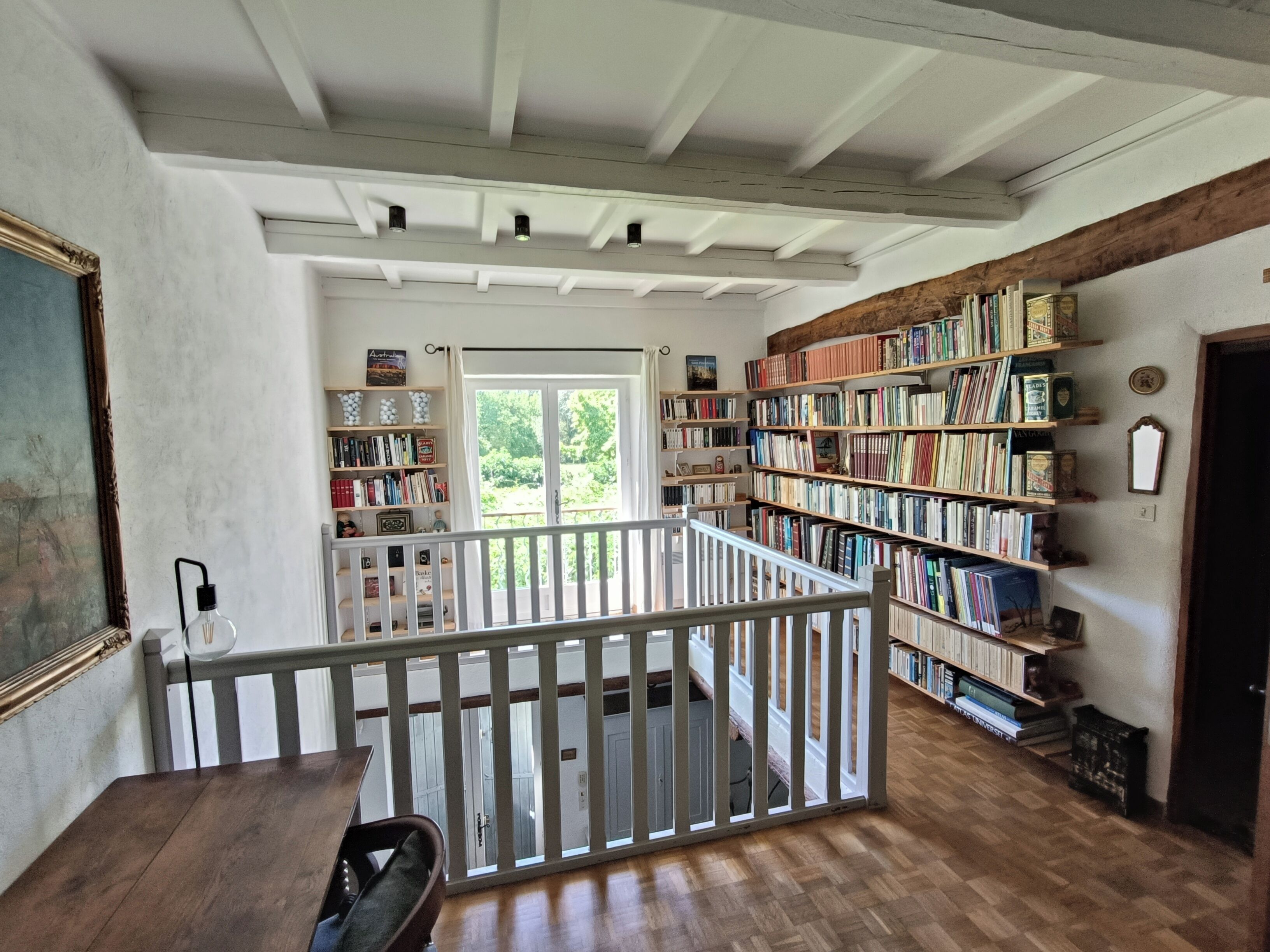 A spacious room with a wooden floor, a white railing, and a bookshelf filled with books.