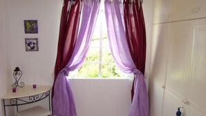 Une pièce avec une fenêtre, des rideaux violets, une petite table avec des tasses, une chaise, un tapis violet et une porte avec un porte-clés.