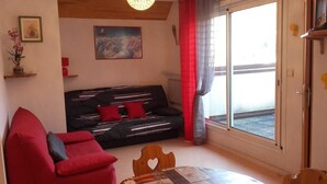 A living room with a black sofa, red cushions, a red armchair, a wooden table, and a large window with red curtains.