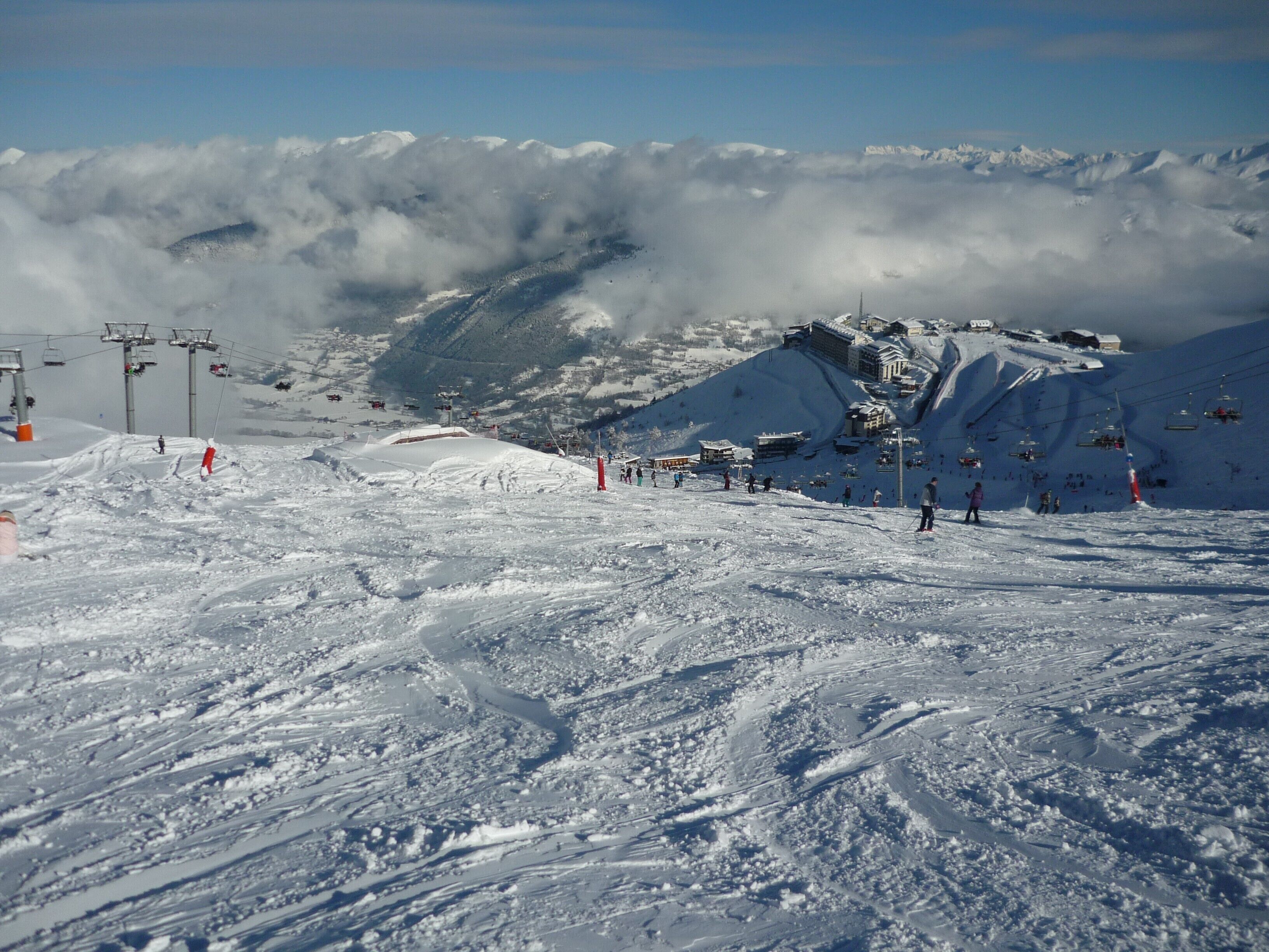 A snow-covered ski slope with ski lifts, a mountain backdrop, and a building complex.