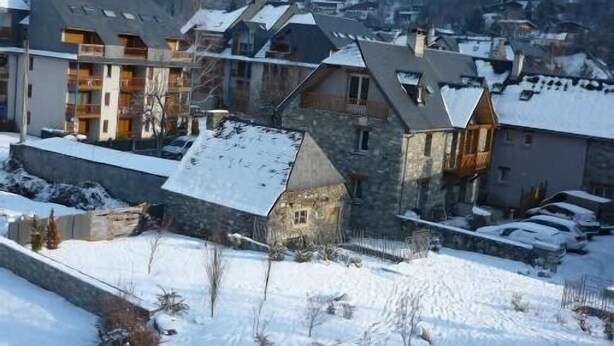 A snow-covered residential area with modern and traditional buildings, including a stone house and a multi-story building with balconies.