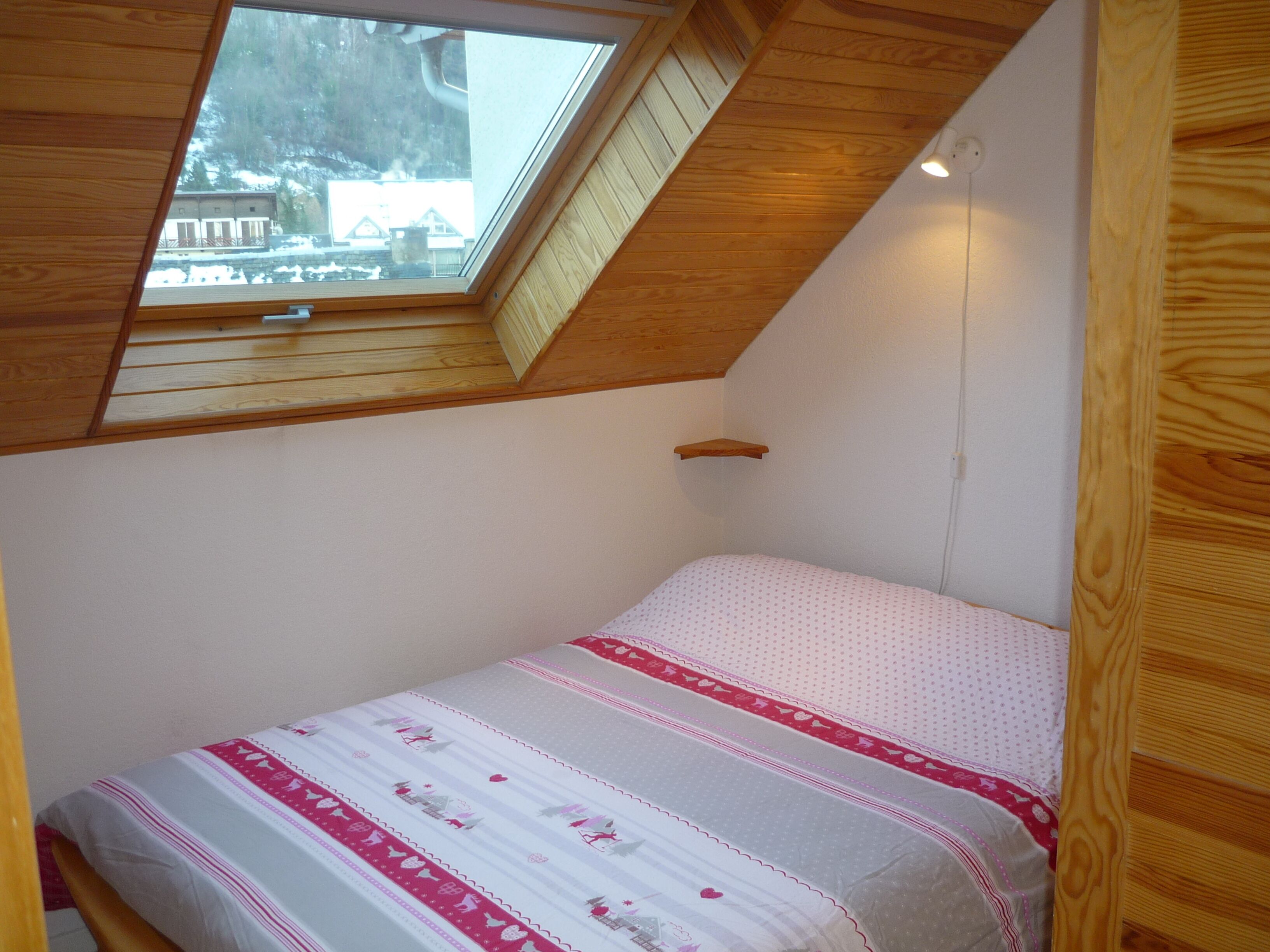 Une chambre avec un plafond en pente et un puits de lumière, comprenant un lit recouvert d’une couvre-lit à motifs et une petite étagère en bois.