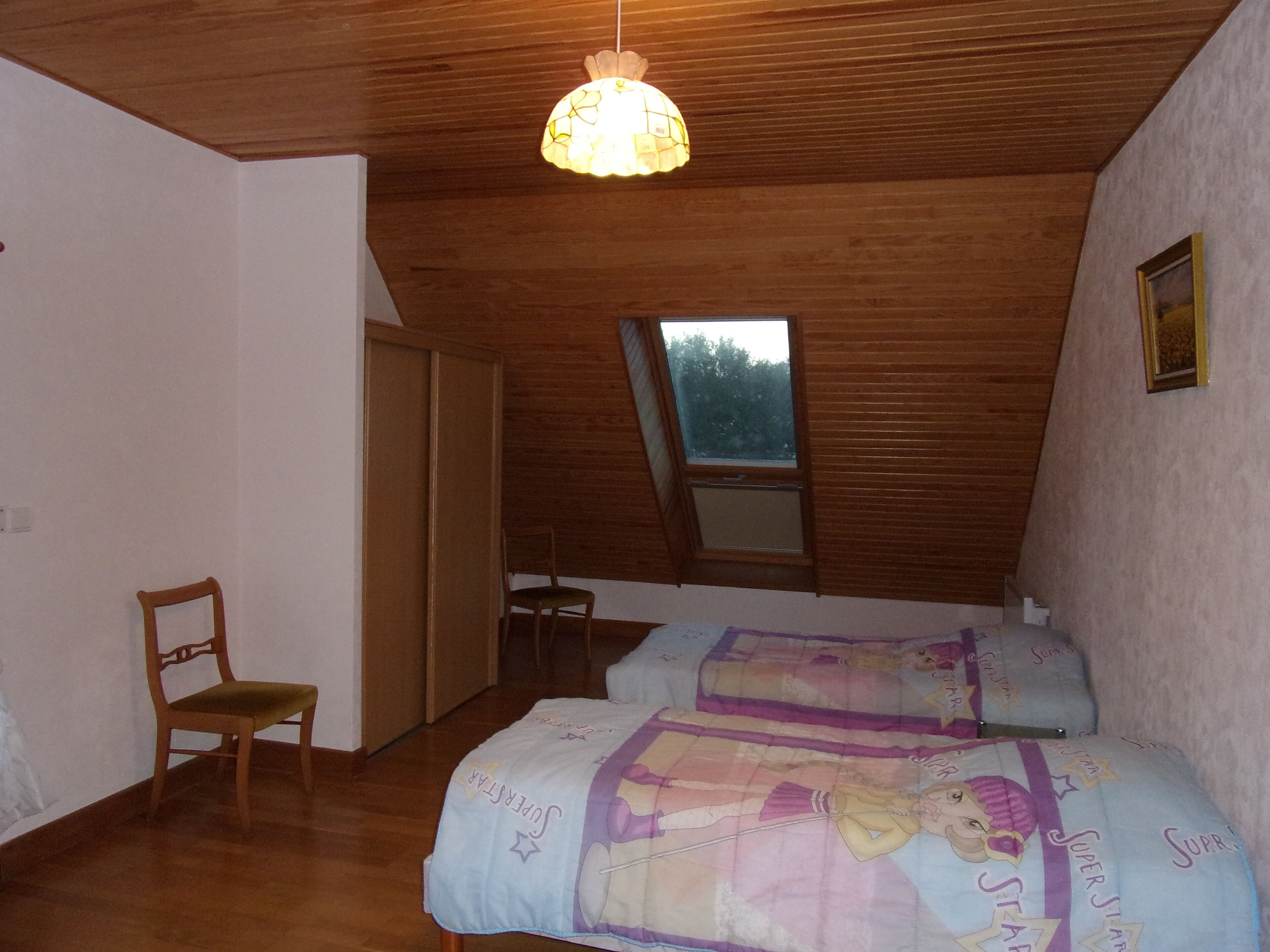 A bedroom with a sloped ceiling, wooden walls, a window, a bed with a Super Star themed bedspread, a chair, and a hanging light fixture.