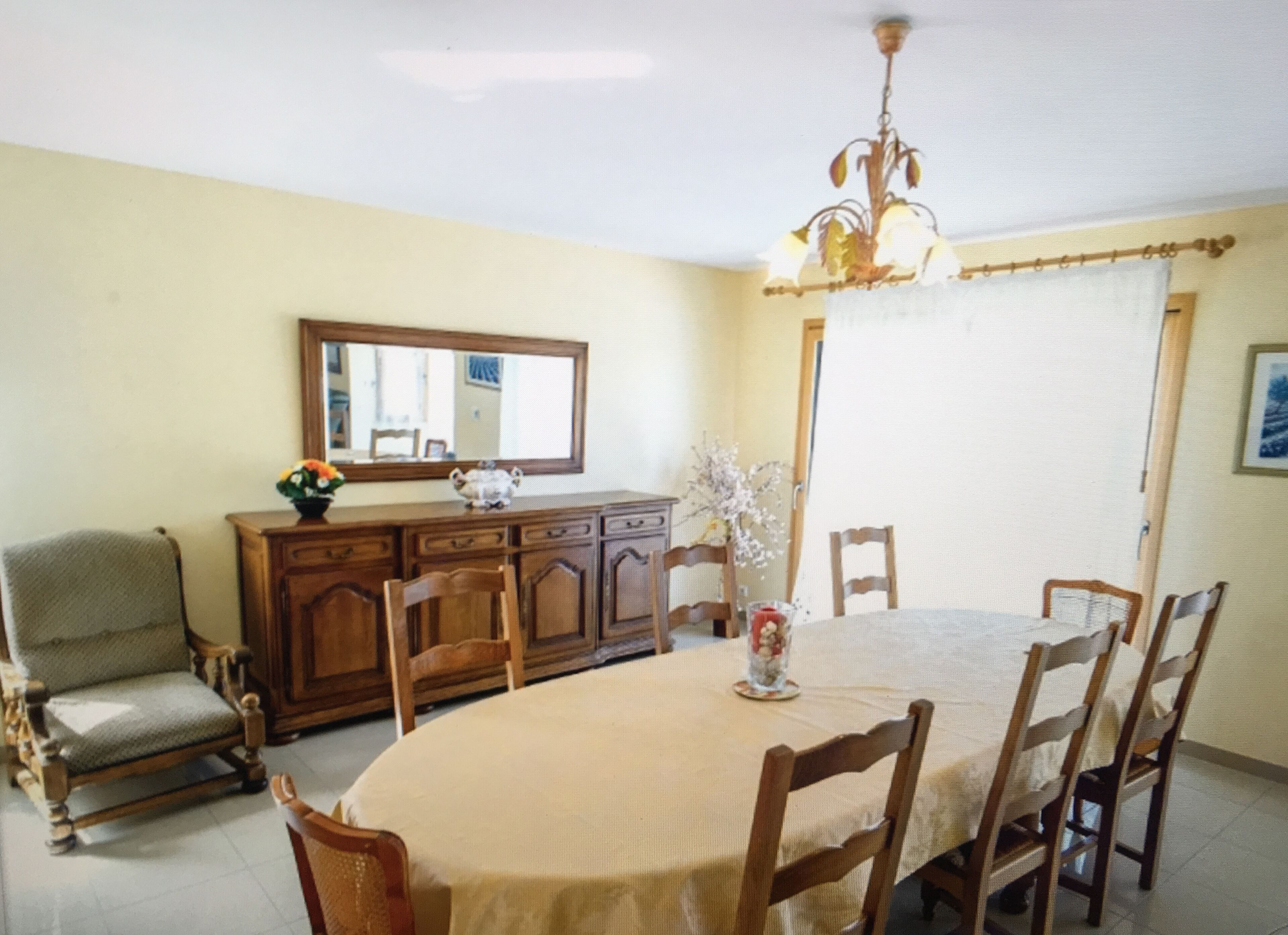 A dining area with a round table, wooden chairs, a chandelier, a mirror, and a cabinet with flowers.