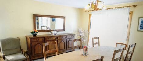 A dining area with a round table, wooden chairs, a chandelier, a mirror, and a cabinet with flowers.
