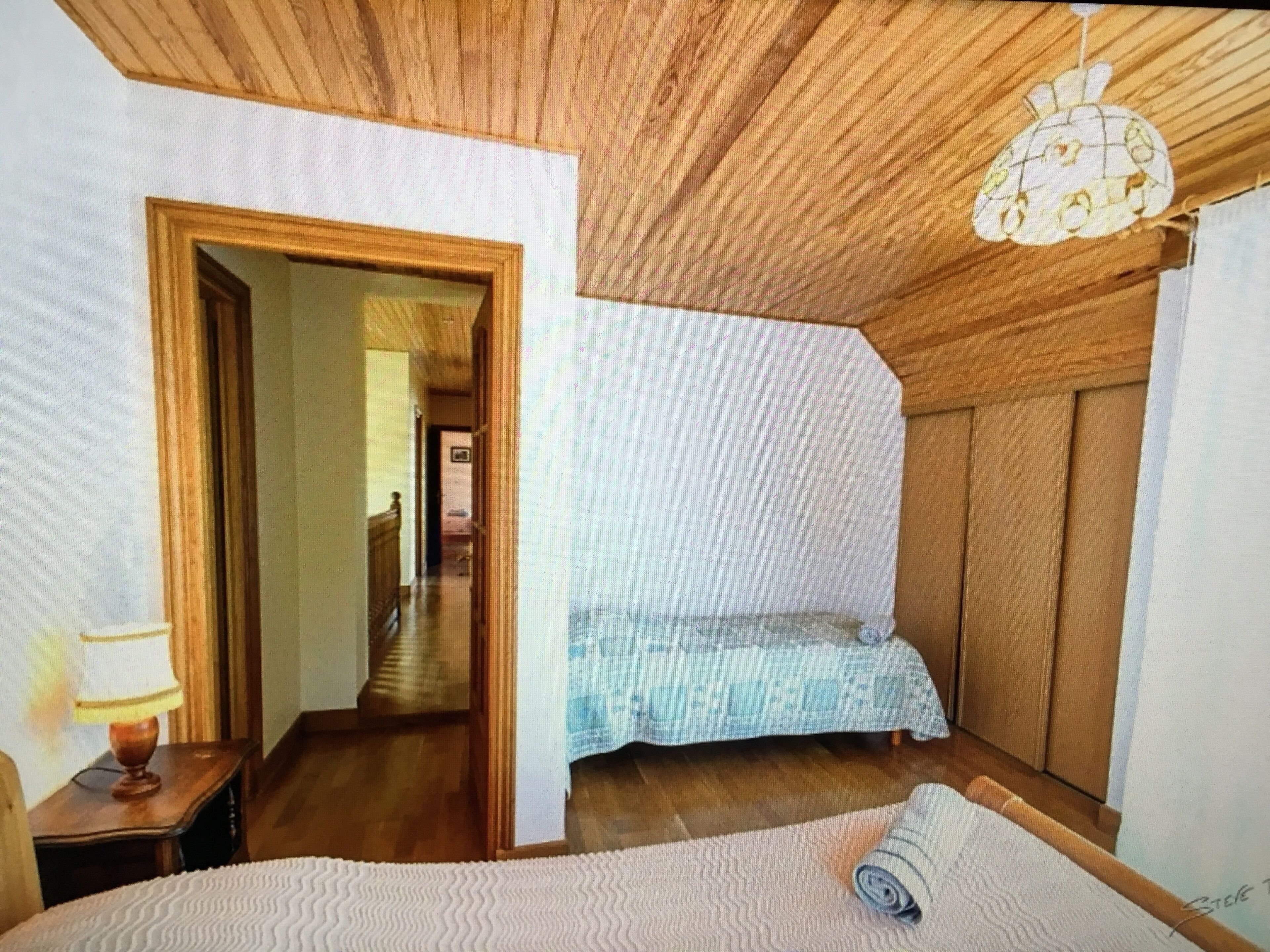 Un dormitorio con techo de madera, una cama con colcha estampada, una mesita de noche con lámpara y un pasillo que conduce a otra habitación.