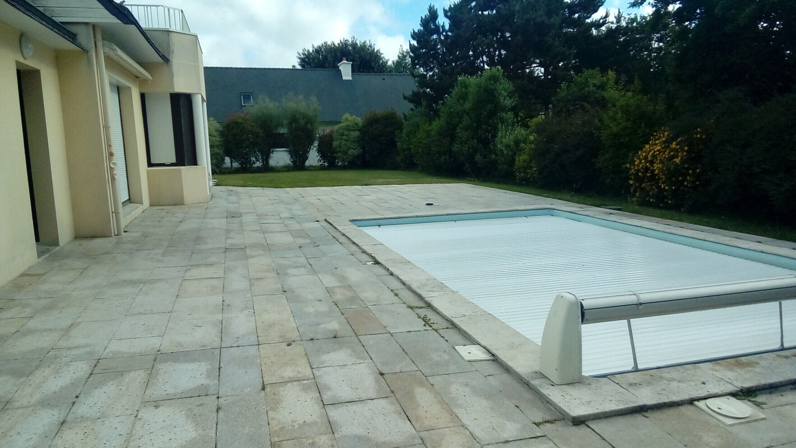 A swimming pool with a tiled deck, surrounded by a garden and a building.