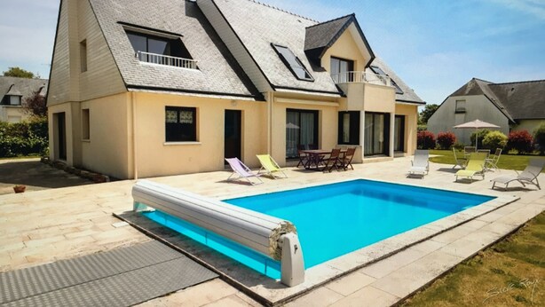 Una casa de dos pisos con piscina, un patio con sillas y un pequeño jardín.