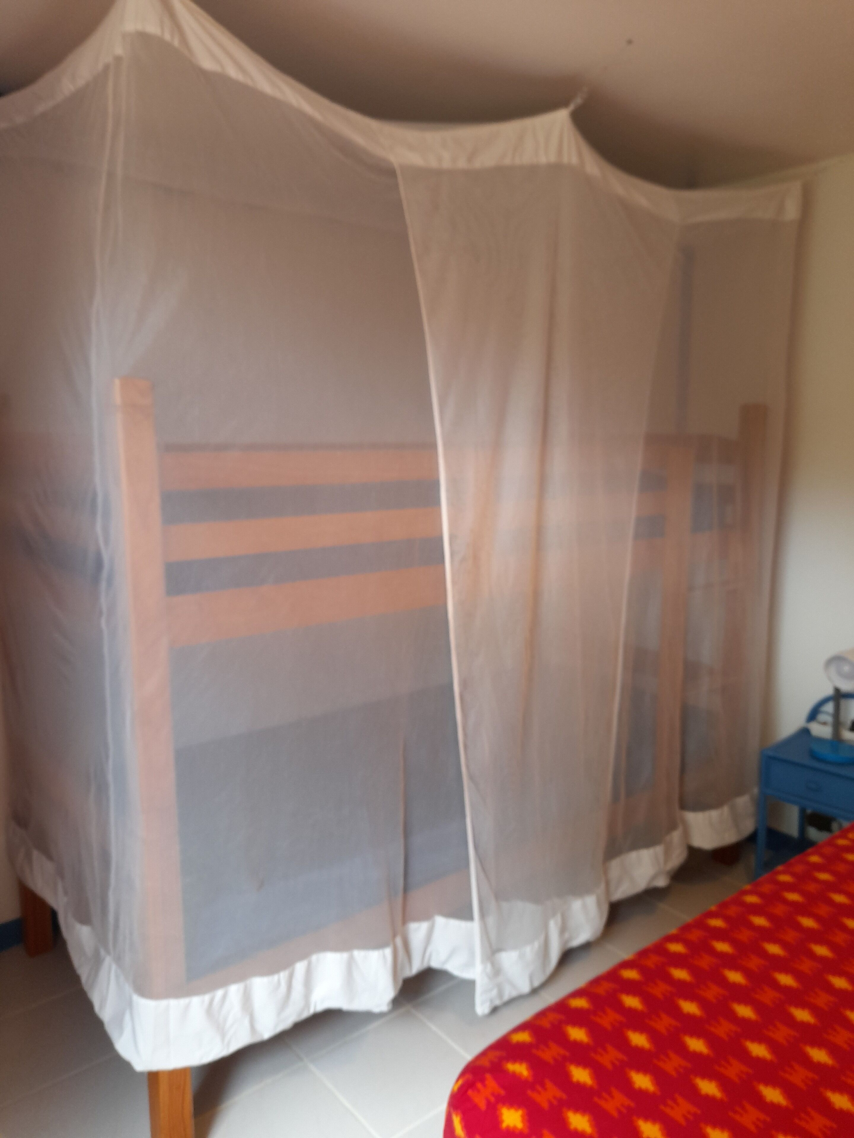 Ein Zimmer mit einem Bett, das mit einem Moskitonetz abgedeckt ist, einem kleinen blauen Tisch mit einer Lampe sowie einer roten, gemusterten Tagesdecke.