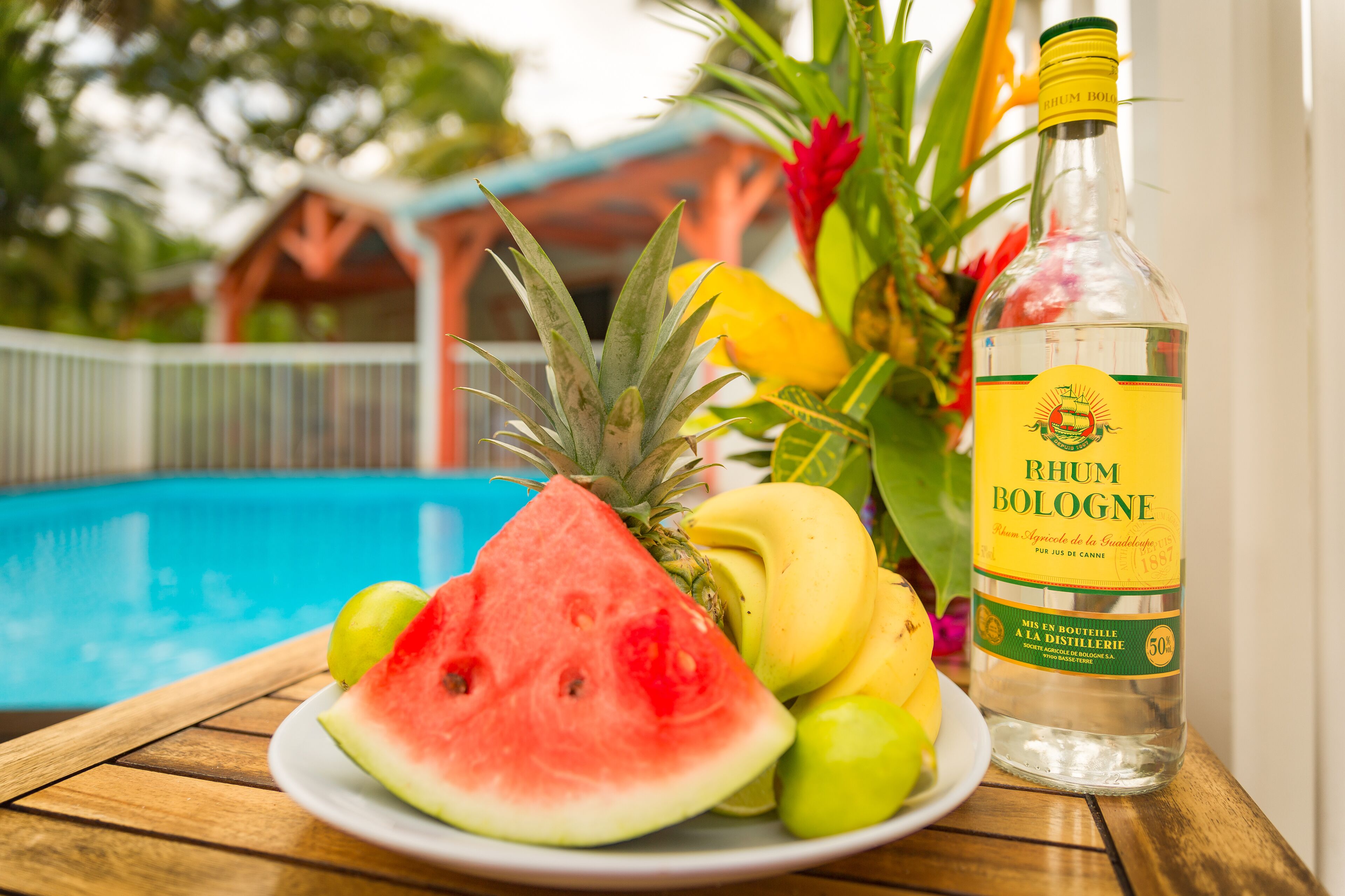 Et bord ved poolen med vandmelon, ananas og bananer, samt en flaske rom.