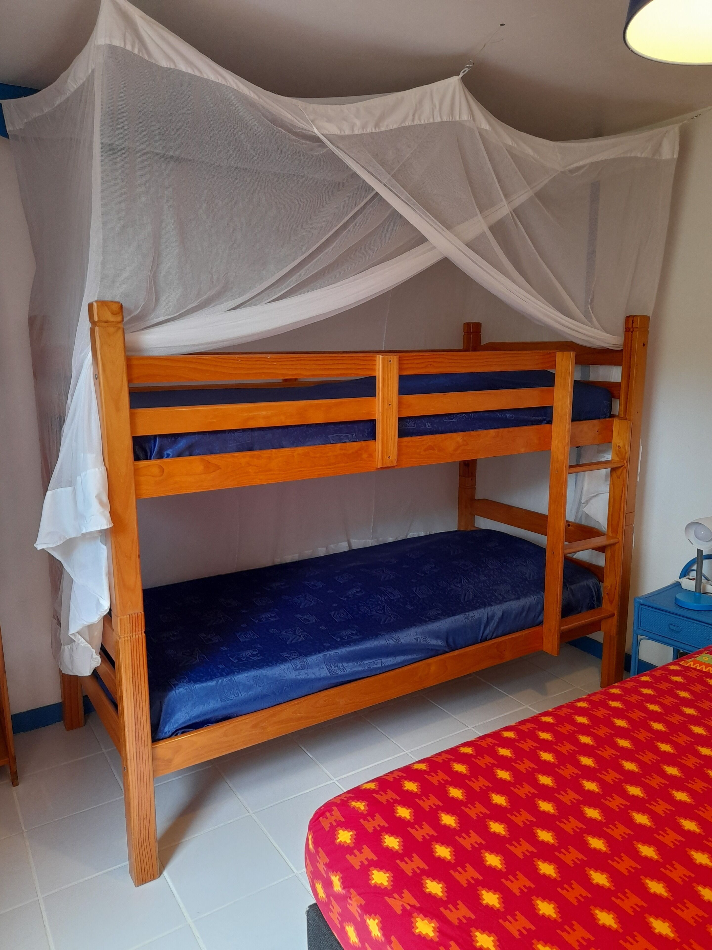 A bunk bed with a mosquito net, a small table with a lamp, and a red patterned bedspread.