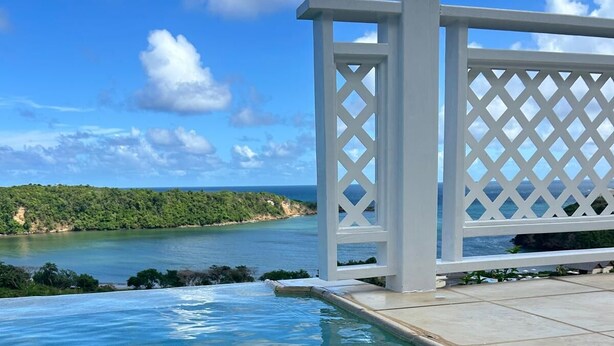Una piscina de blau clar amb una tanca de lletja blanca i vista a l'oceà i al verd.