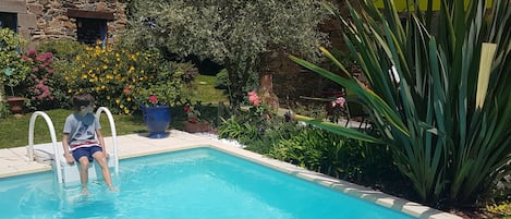 Uma piscina com uma casa de pedra ao fundo, rodeada por diversas plantas e flores.