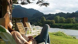 A person relaxing on a chair by a riverside with a view of mountains in the background.