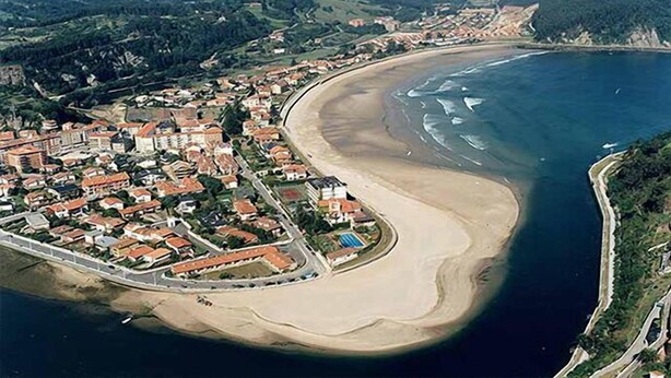 An aerial view of a coastal town with a beach, houses, and a road leading to the shore.