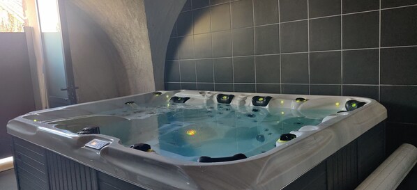 A hot tub with a control panel, surrounded by dark tiles and a stone wall.