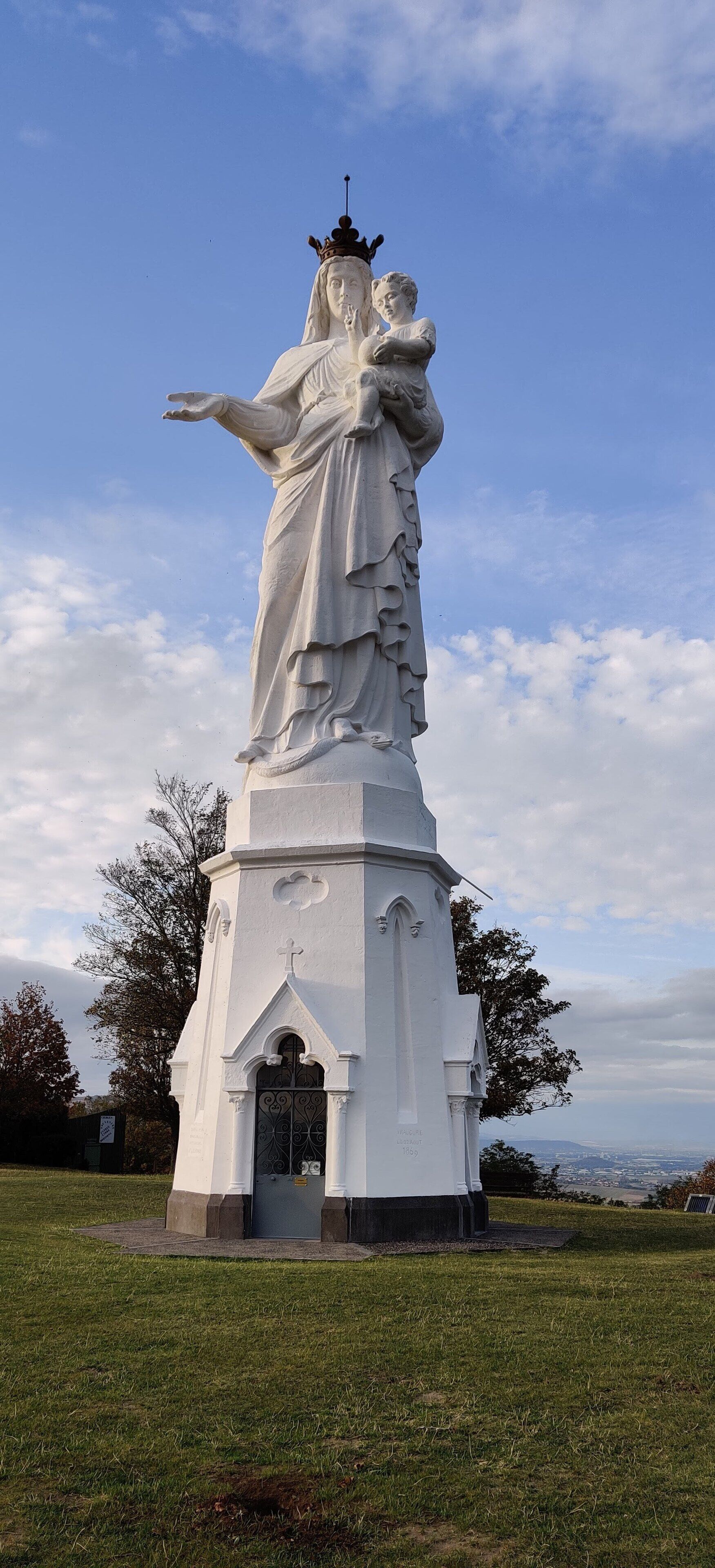 A white statue of a woman holding a child, standing on a pedestal with a cross, set against a clear sky.