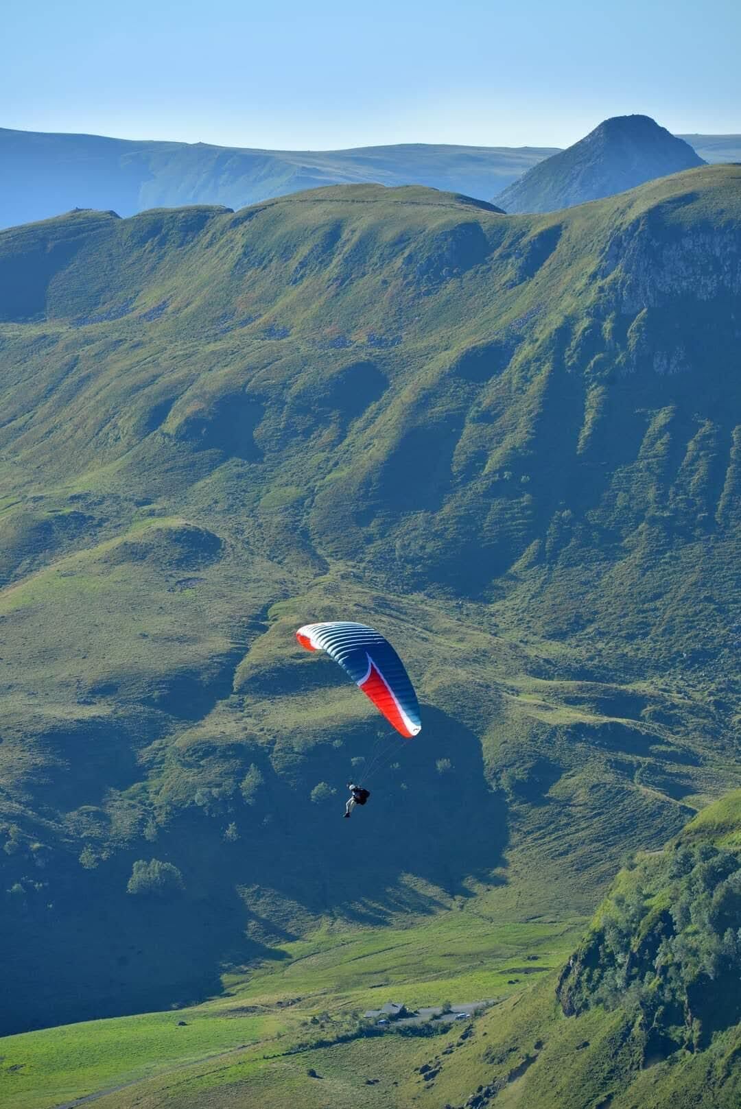 En person paraglider over et bjergrigt landskab med grønne bakker og en klar himmel.