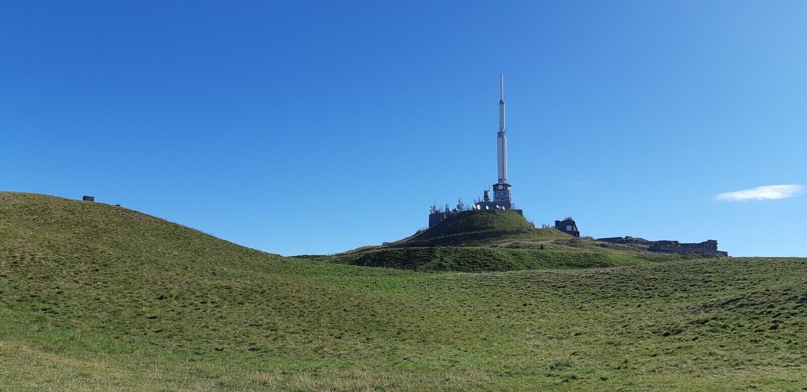 Ein schlanker, hoher Turm auf einer grünen Hügelkuppe unter blauem Himmel.