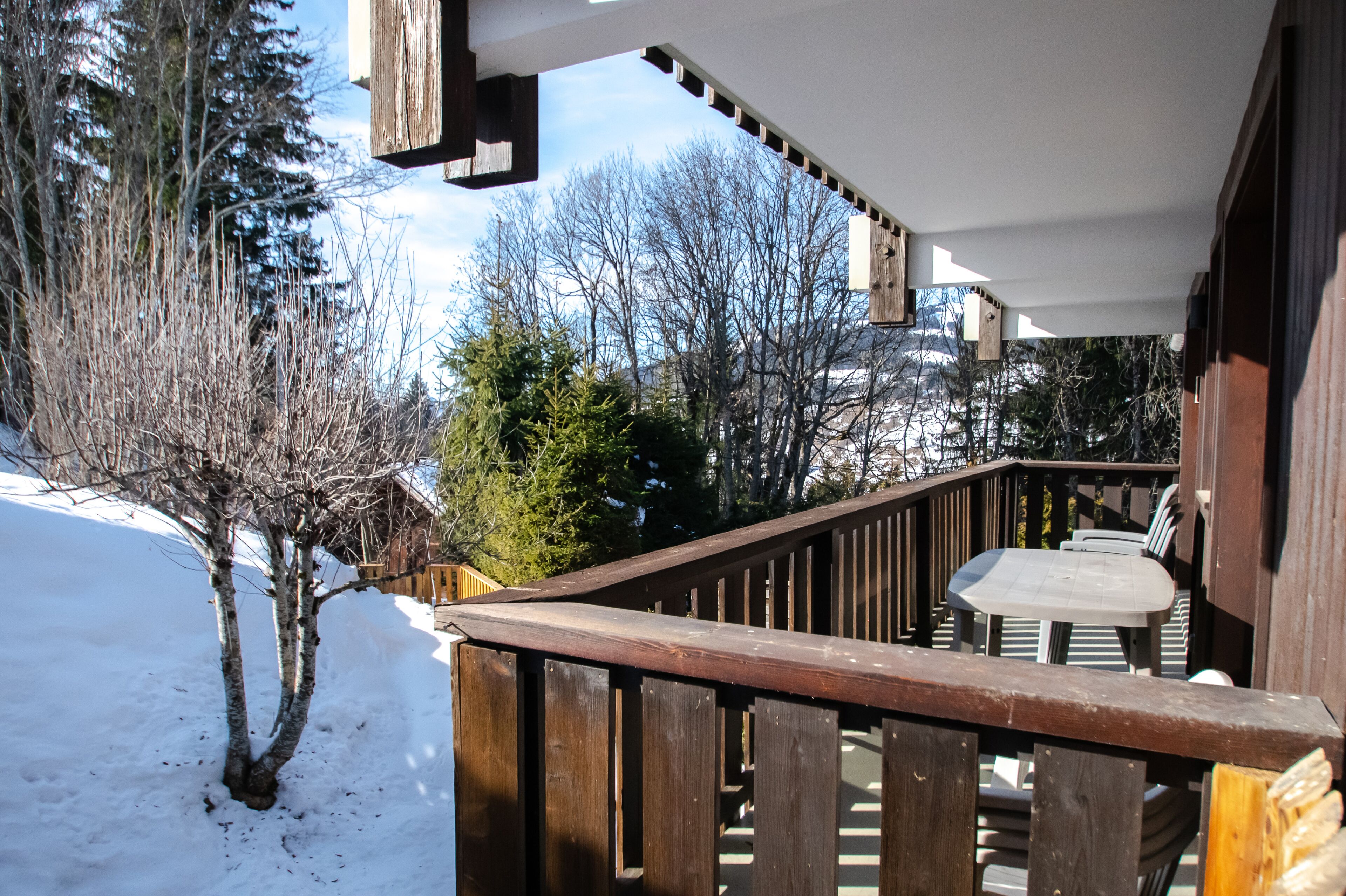 A wooden deck with a table and chairs, surrounded by snow and trees.
