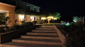 A nighttime view of a building with a staircase leading up to a door, surrounded by well-lit landscaping and a pool area.