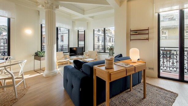 A modern living room with a sofa, a coffee table, and a TV. There is a balcony with a railing and a view of buildings. - Allegra by FeelFree Rentals (Donostia)