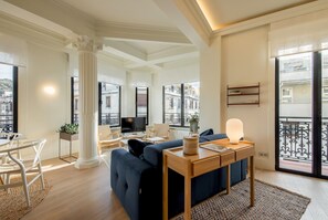 A modern living room with a sofa, a coffee table, and a TV. There is a balcony with a railing and a view of buildings.