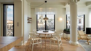 A modern living room with a round wooden table, white chairs, a large window with a view of buildings, and a decorative column. - Allegra by FeelFree Rentals (Donostia)