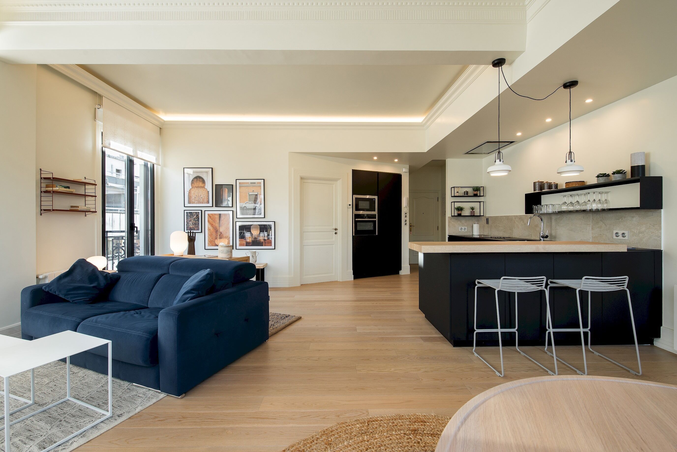 Una sala moderna con un sofá azul oscuro, una mesa de centro blanca y una cocina con taburetes altos y una encimera.