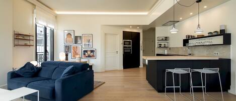 Una sala moderna con un sofá azul oscuro, una mesa de centro blanca y una cocina con taburetes altos y una encimera.