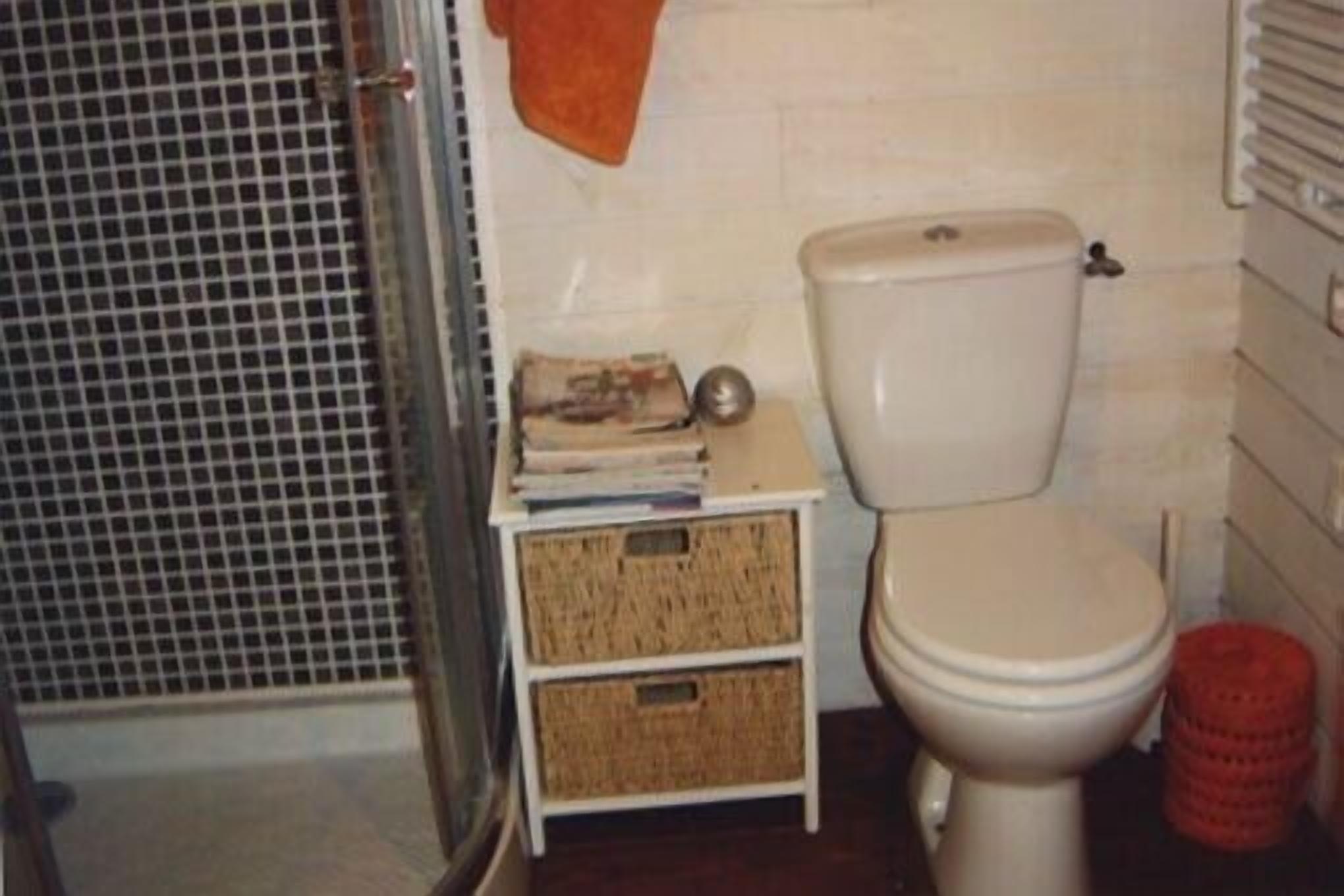 Une salle de bain avec des toilettes, une petite étagère blanche avec des paniers, une poubelle rouge et un espace de douche carrelé.