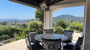 Eine überdachte Terrasse im Freien mit einem runden Tisch und Stühlen, einer Pendelleuchte und Blick auf eine hügelige Landschaft.