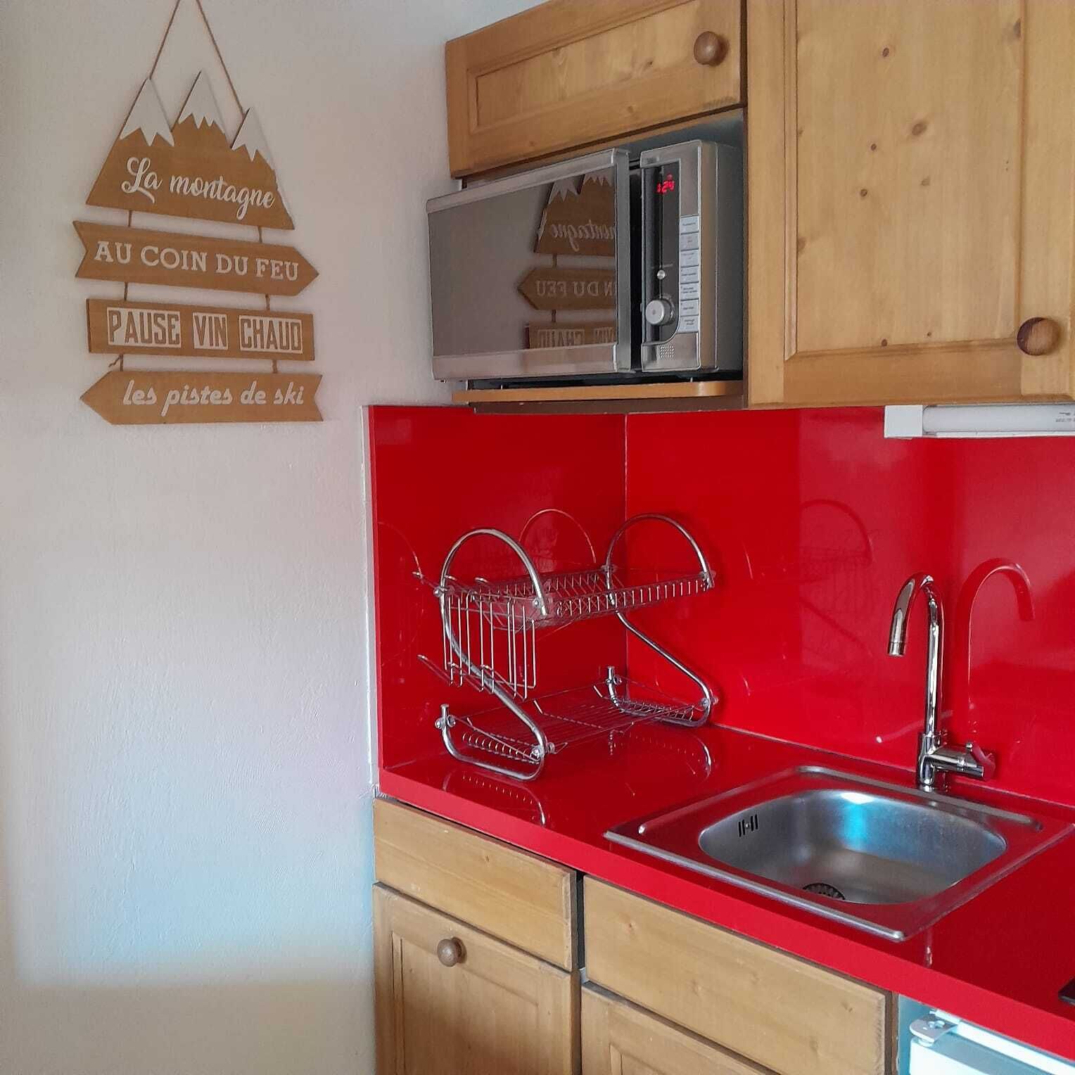 A kitchen with a red countertop, a stainless steel sink, and wooden cabinets. There is a microwave on the countertop and a wooden sign with text on the wall.