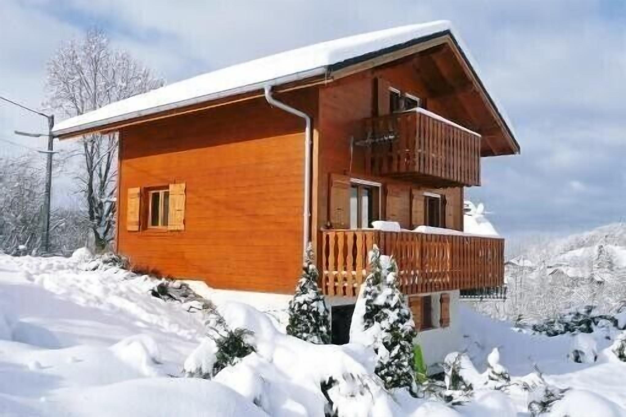 A two-story wooden cabin with a balcony, surrounded by snow-covered trees and mountains.