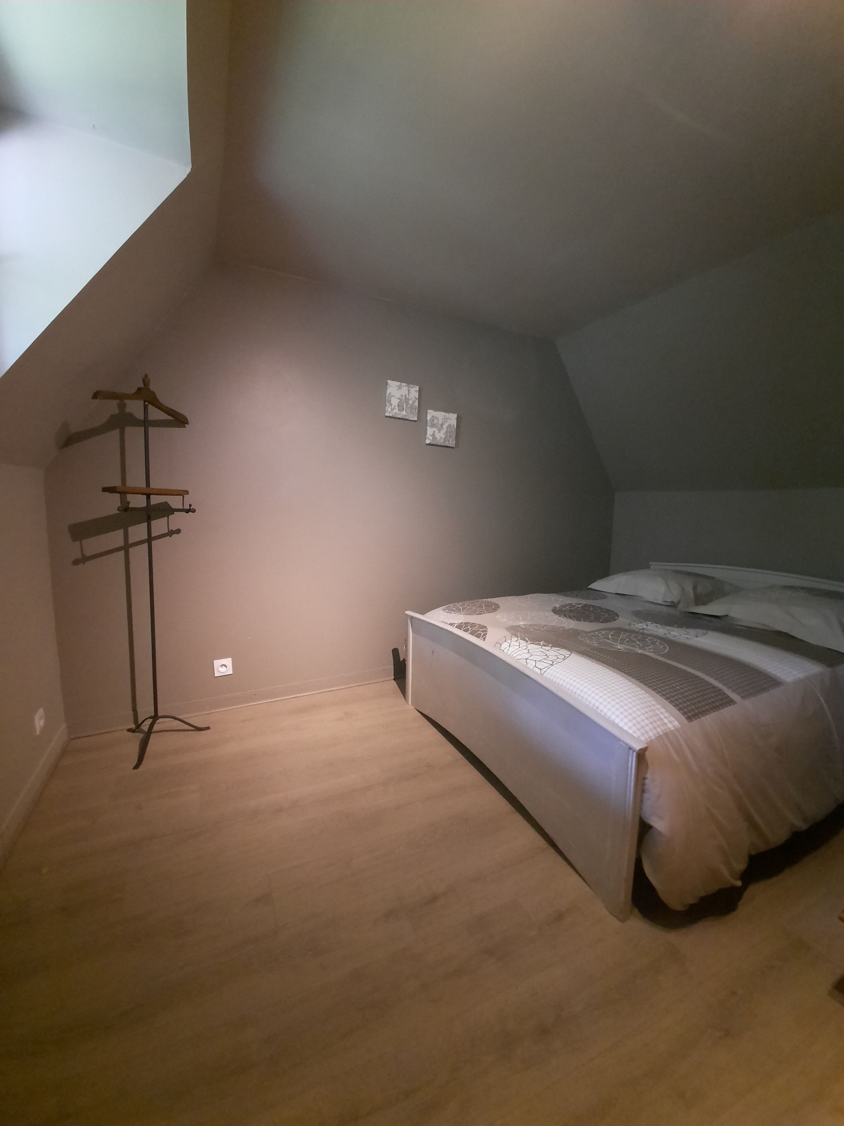 A bedroom with a sloped ceiling, a wooden floor, a bed with patterned bedding, a coat rack, and two small wall-mounted items.