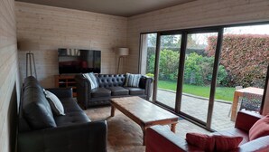 A modern living room with a black leather sofa, a red sofa, and a wooden coffee table. There is a television on a stand and a floor lamp. The room has large windows with a view of a garden. - In the estuary of the Seine, a few minutes away from Honfleur, a real Longere (Eure)