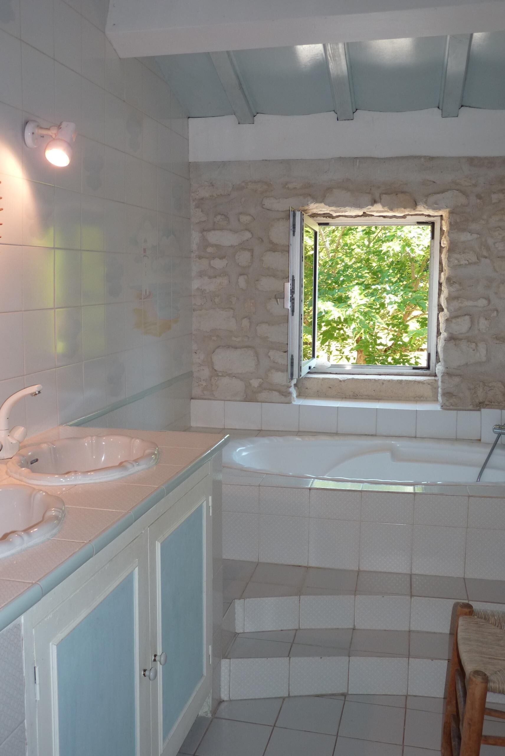 A bathroom with a stone wall, a window with a view of greenery, a bathtub, and a sink with a mirror.
