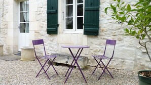 Un bâtiment en pierre doté de deux fenêtres, chacune ornée de volets verts. Devant les fenêtres, on aperçoit une petite table et deux chaises violettes.