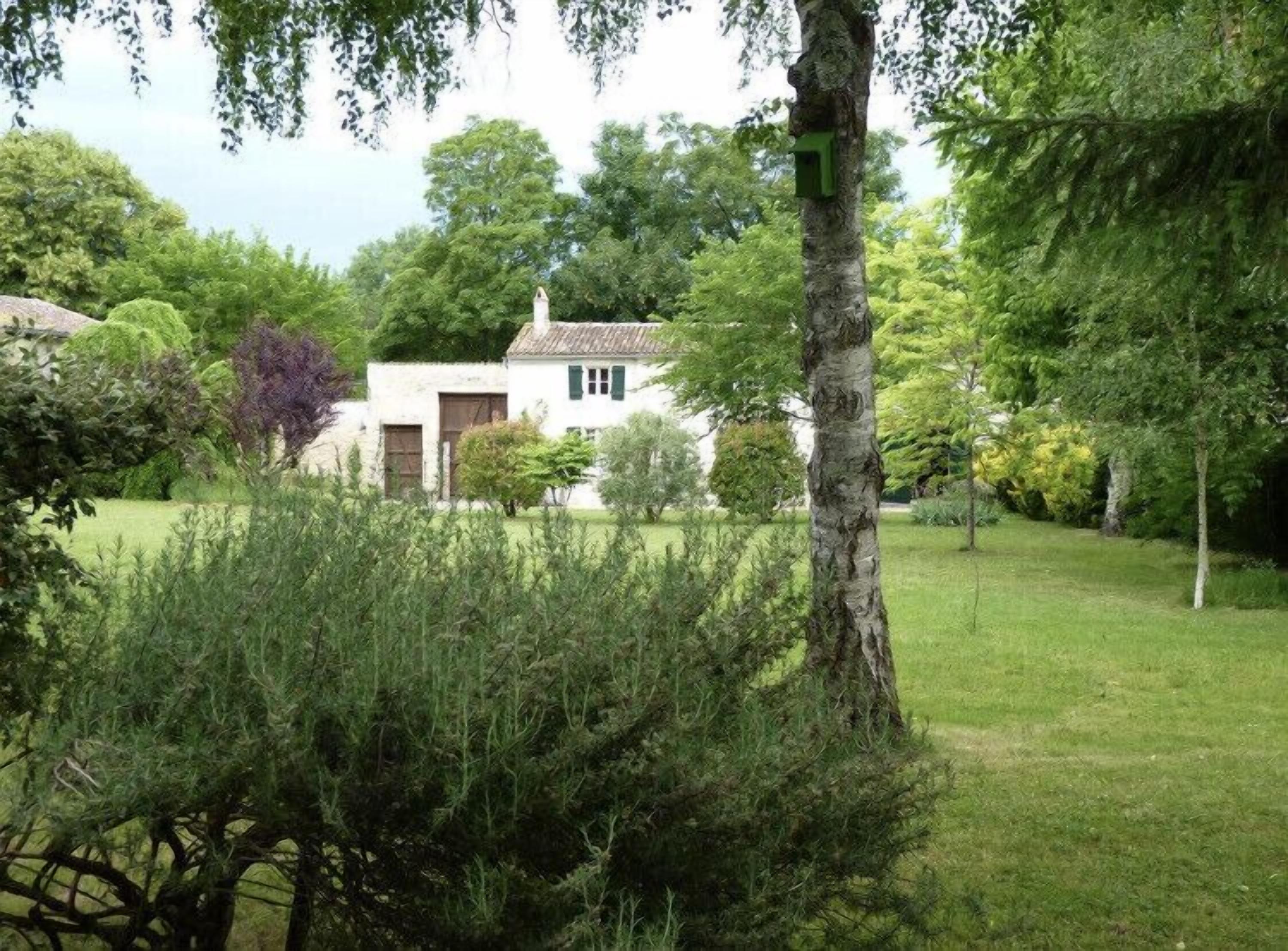 Una casa bianca con una porta verde e finestre, circondata da vegetazione rigogliosa e alberi.
