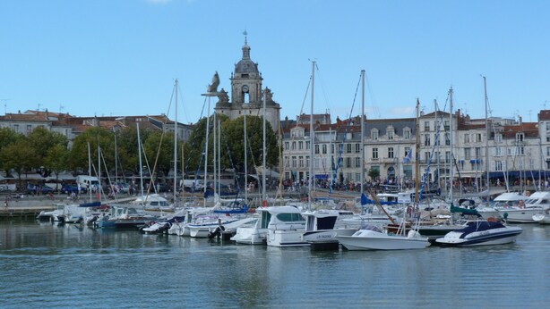 Un port de plaisance où sont amarrés de nombreux bateaux, un bâtiment historique avec un clocher en arrière-plan et un ciel dégagé.