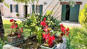 Un bâtiment en pierre avec des volets verts, entouré d’une végétation luxuriante et de plantes en fleurs.