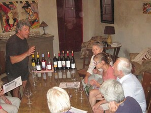 A group of people seated around a table with multiple bottles of wine and glasses, in a room with a wooden table and chairs, a lamp, and a door in the background.