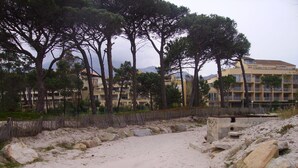 Un sentier de plage sablonneux bordé d’une clôture en bois, de gros rochers et de grands pins. Au fond, on aperçoit des immeubles à plusieurs étages dotés de balcons.