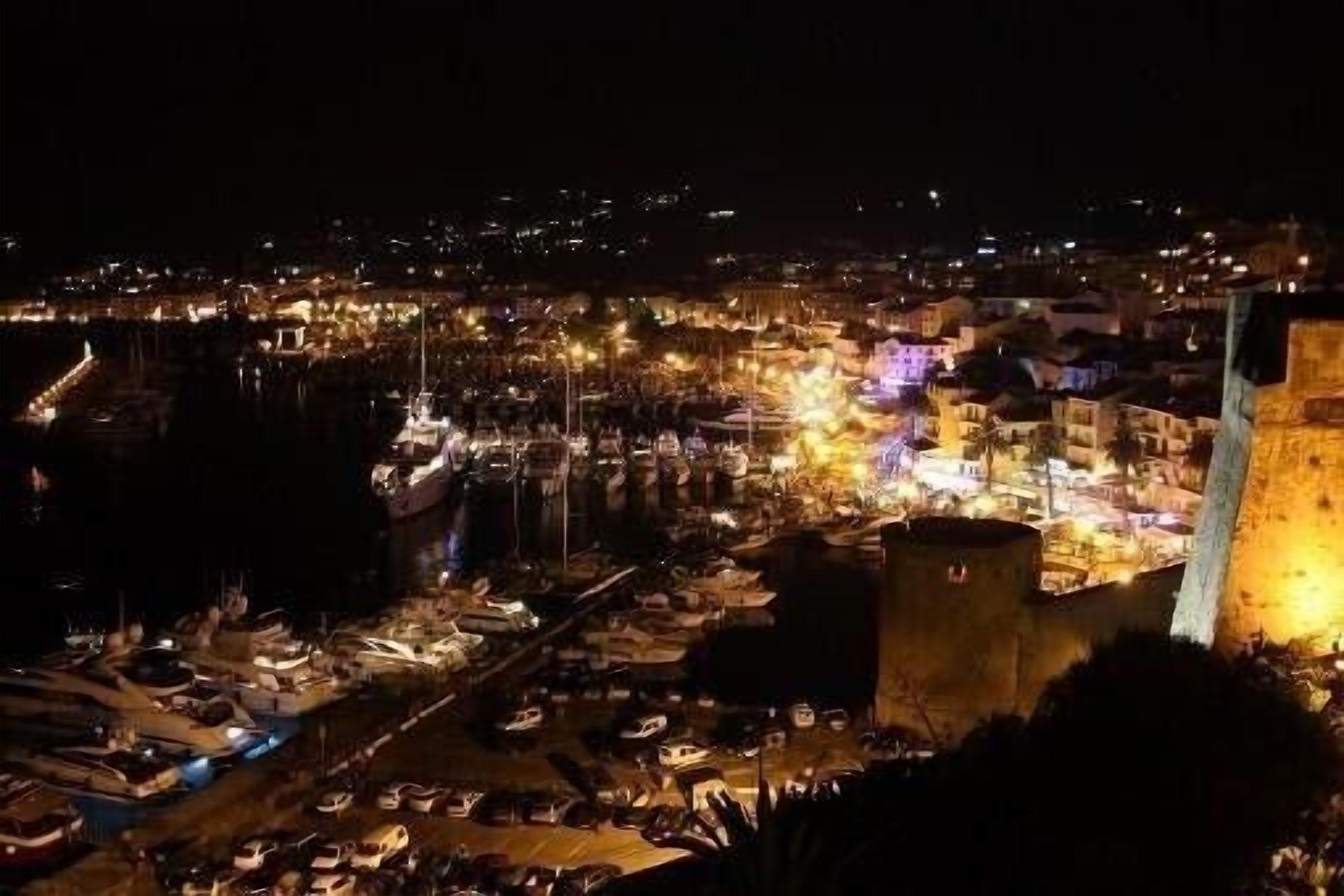 A nighttime view of a coastal town with a prominent tower and a marina filled with boats.