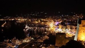 Une vue nocturne d’une ville côtière, avec un imposant beffroi et un port de plaisance rempli de bateaux.