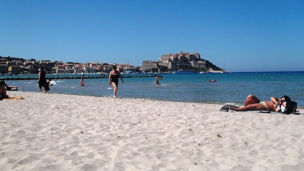 Une plage où des gens se prélassent au soleil et se baignent, un château en arrière-plan et des bateaux sur l’eau.