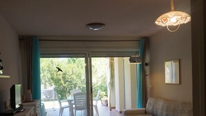 Un salon comprenant un canapé, un pouf et un coin repas avec des chaises et une table. Une porte-fenêtre coulissante donne accès à un espace extérieur aménagé avec une table et des chaises.