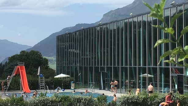 Zona de piscina con un edificio de cristal, un tobogán y montañas al fondo.