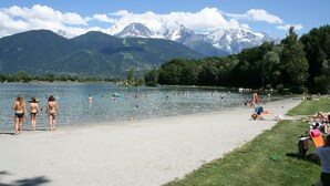 Una playa junto al lago, con gente nadando y tomando el sol, rodeada de montañas y árboles.