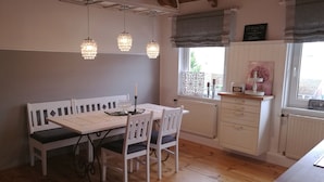 Uma cozinha acolhedora com mesa de madeira, cadeiras brancas, luminárias pendentes e uma janela com vista.